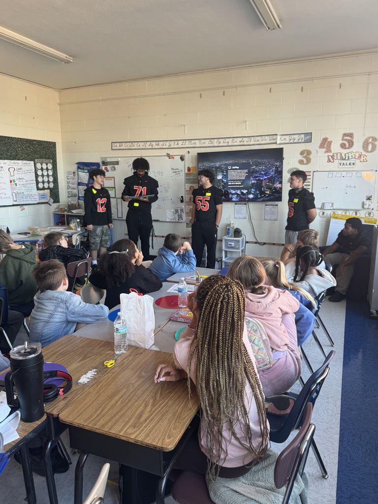 four south charleston high school football players standing in front of an elementary classroom reading to students.