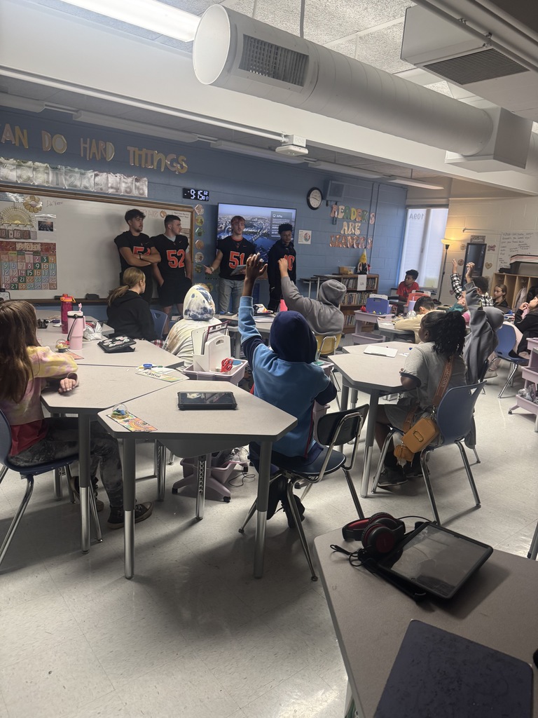 four south charleston high school football players standing in front of an elementary classroom reading to students.