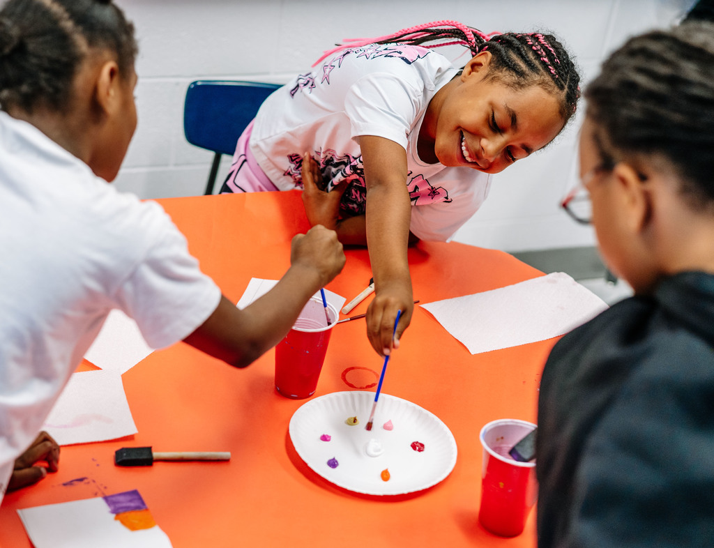 three girls laughing while reaching for paint with their paint brush.