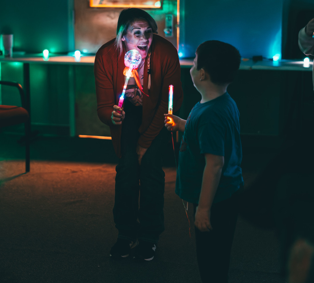 a student and a teacher standing in a dark room holding glow wands and dancing.