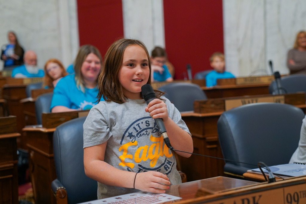 a student standing and speaking into a microphone at a delegates desk in the west virginia house of delegates chamber.