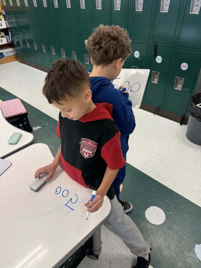 two students are standing back-to-back, one student is facing his desk and the other student is facing a row of lockers, and they are writing down their answers for a math problem during an interactive math lesson.