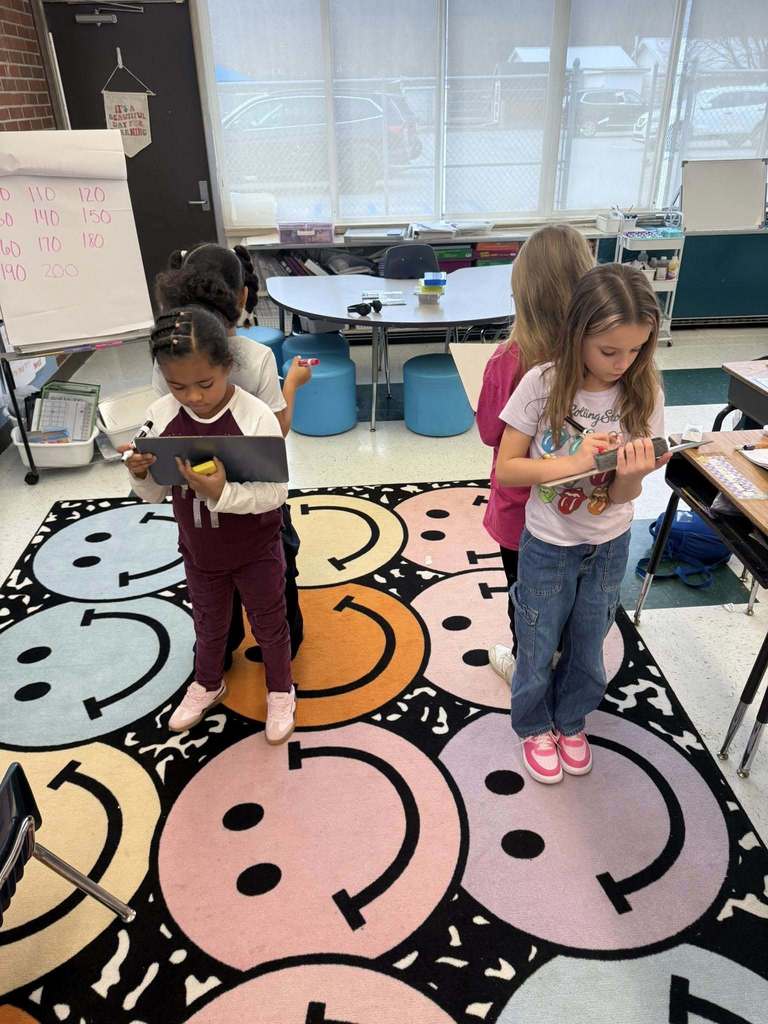 Four students, broken up into pairs of twos and standing with their backs against their partner, writing on a small white board during an interactive math lesson.