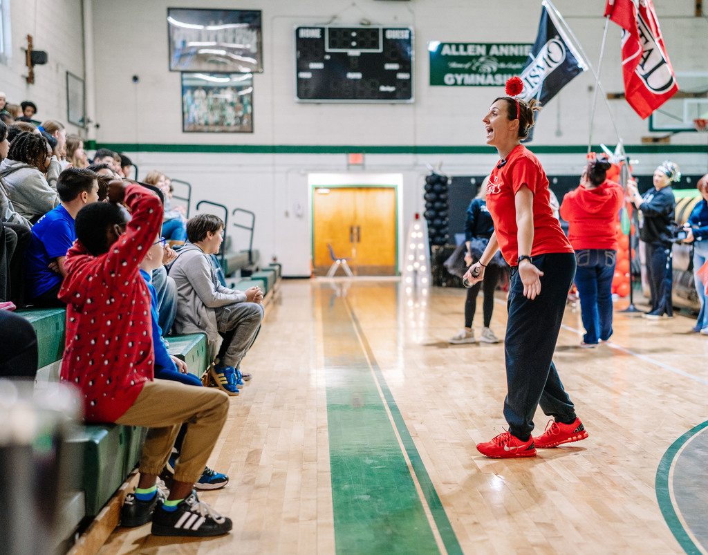 Horace Mann Middle School's vice principal standing in front of the bleachers, where the students are sitting, encouraging them to get excited. Teachers in the background are waving flags for the event.