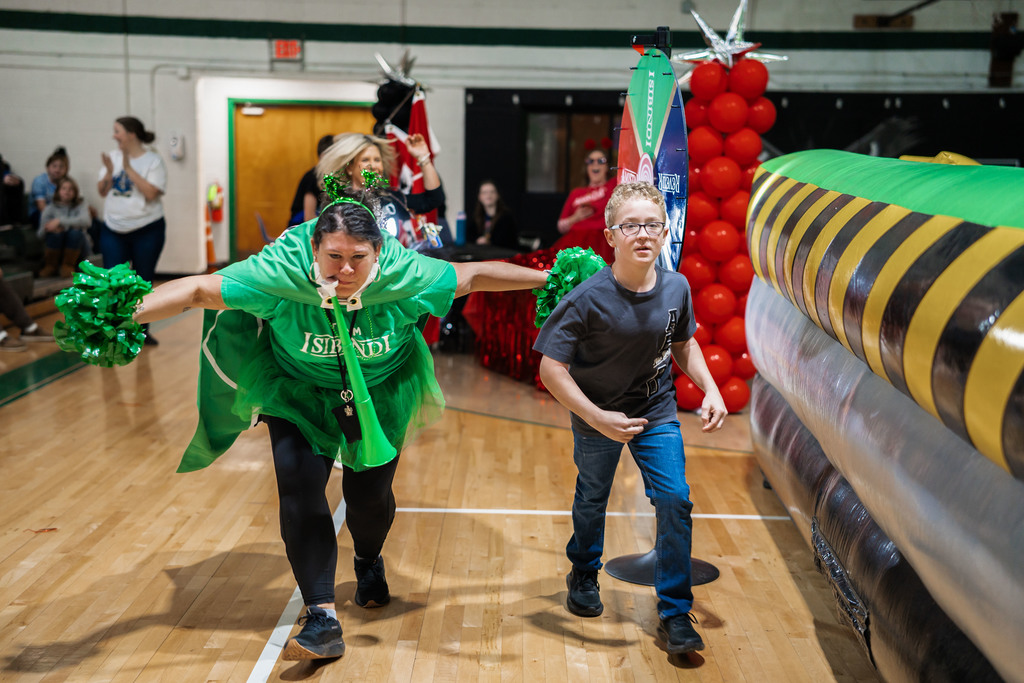 A student and a teacher, who is blowing an air horn, wearing a flag, and holding pom poms, are running down the gym floor after the student found out he was selected in to the green house.