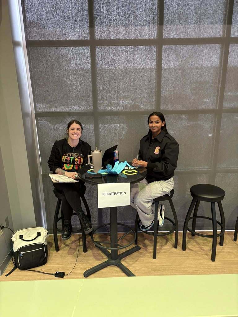 Two members of the Capital High School JROTC smiling for a picture while sitting at the registration table for a food giveaway for families during the holidays. 