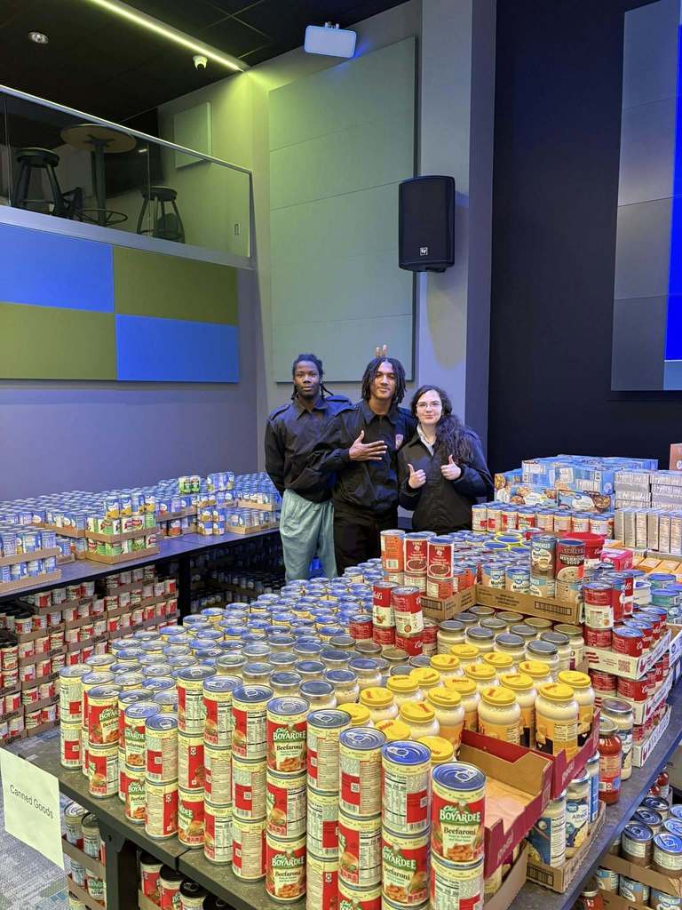 Three Capital High School JROTC students posing for a picture behind a table stacked with a variety of canned food.