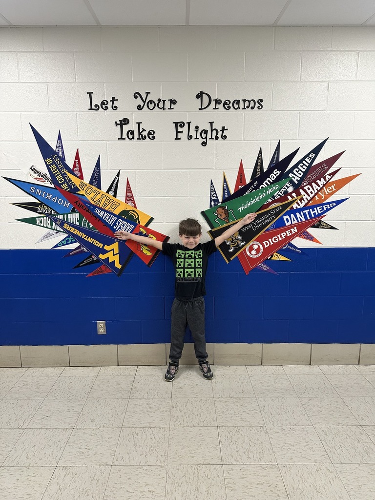 A blue and white wall with flags of universities on it arranged to look like wings. Above the flags stylized letters spell out "Let Your Dreams Take Flight." A young student is standing in front of the wall smiling with his arms stretched out wide.