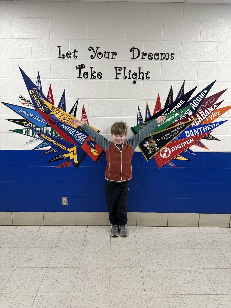 A blue and white wall with flags of universities on it arranged to look like wings. Above the flags stylized letters spell out "Let Your Dreams Take Flight." A young student is standing in front of the wall smiling with his arms stretched out wide.