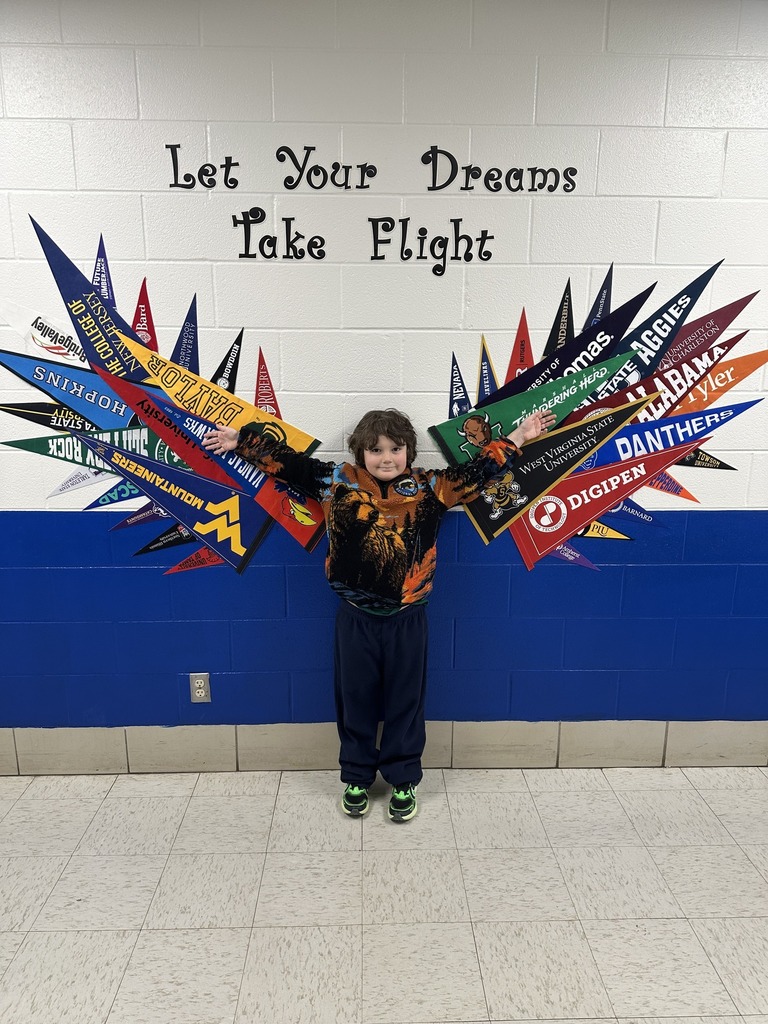 A blue and white wall with flags of universities on it arranged to look like wings. Above the flags stylized letters spell out "Let Your Dreams Take Flight." A young student is standing in front of the wall smiling with his arms stretched out wide.