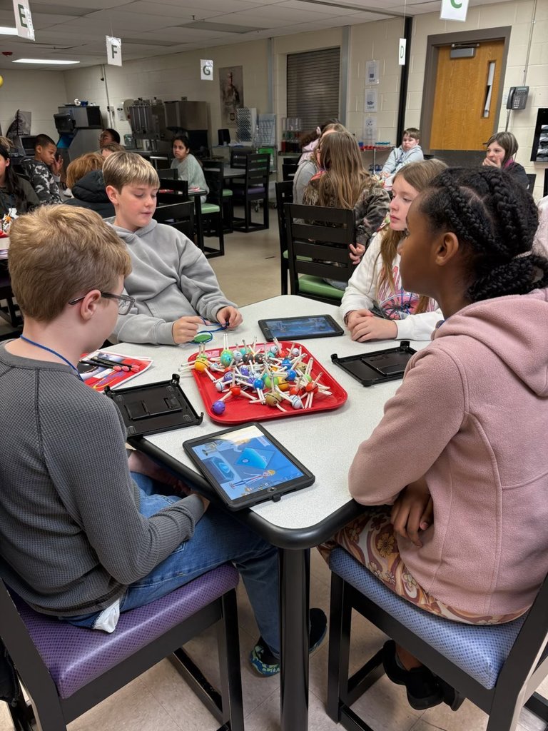 Four students sitting at a table getting ready to do an experiment. Two students have iPads. In the center of the table there is a red serving tray that has molecule figures on top of it.