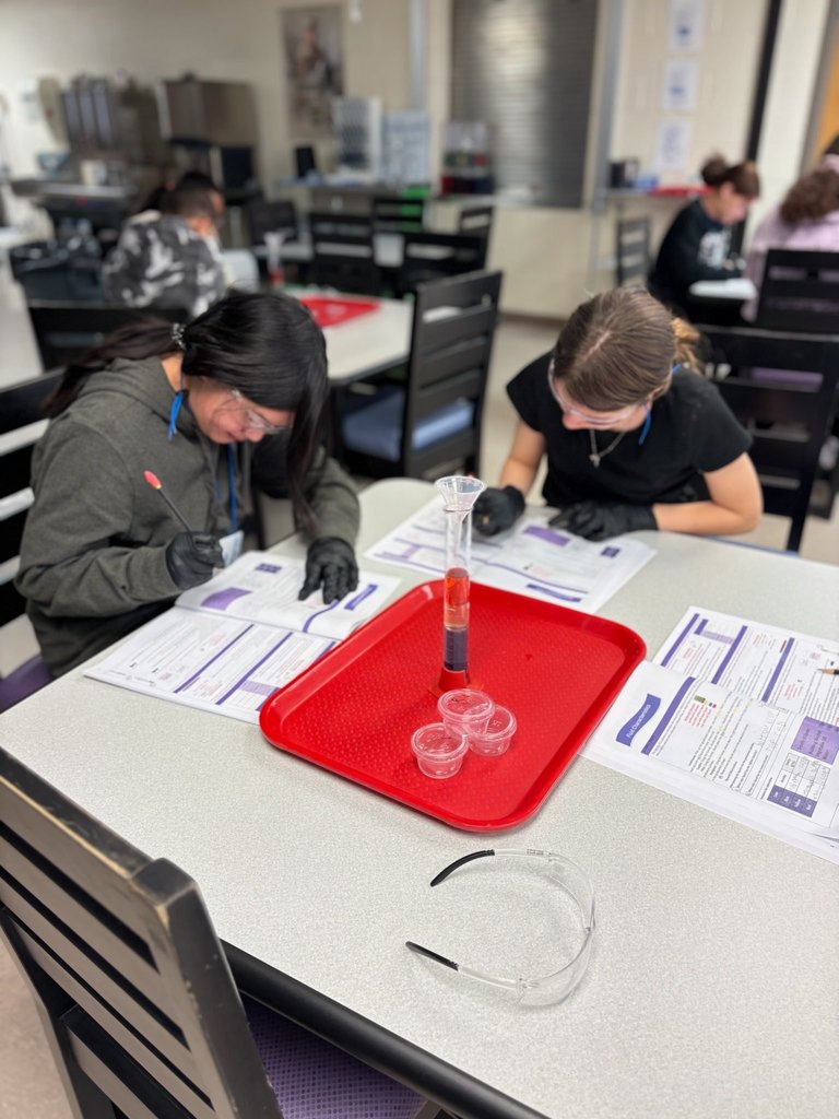 Two students, slightly out of focus, sitting at a table with black lab gloves on writing in their lab notebook. Just in front of them is a clear cylinder filled halfway with different liquids. A pair of lab glasses and three small, empty, plastic containers are sitting in front of the cylinder.