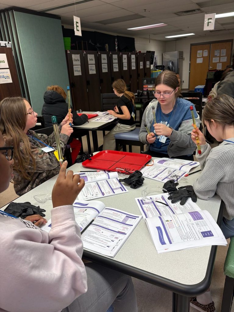 Four students sitting at a table, holding up a thin, long, and clear tube that has liquid in it. There are four lab notebooks on the table along with their lab gloves and goggles.