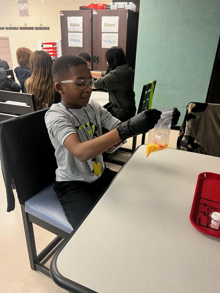 A student sitting at a table holding a plastic baggie that is a quarter of the way filled with a yellow substance. The student is wearing lab gloves and googles and has an excited look on his face.