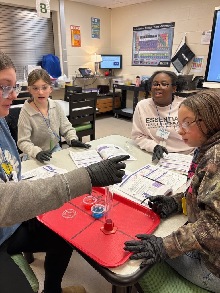 Three students are watching an employee at Starbase drop something into a clear glass cylinder. All three students are looking intently at the cylinder.