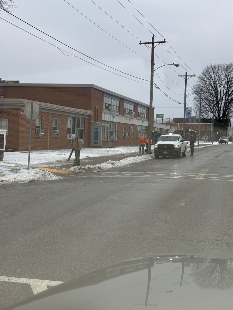 In the foreground, a city of South Charleston worker is using a leaf blower to blow snow off of the main sidewalk at South Charleston Middle School. Behind that man, about halfway down the sidewalk, two men are using snow shovels to clear snow from the sidewalk at the school. Beside the two men with the snow shovels, a city work truck is parked and two men are getting snow removal equipment out of the bed of the truck. 