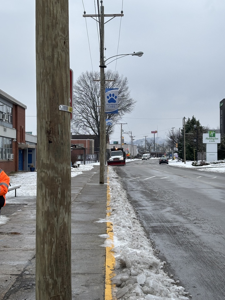 A city of south charleston snow plow is driving down the street in front of South Charleston Middle School. The snow plow is about to begin clearing a strip of snow that is just in front of the sidewalk at South Charleston Middle School.