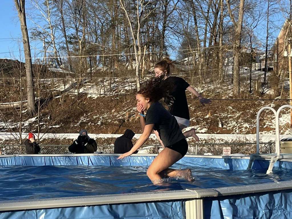 Two girls wearing a tee shirt and shorts jumping into an above ground pool. The legs of the girl closes to the camera have just entered the water. The second girl is still in mid air. In the background a man is taking pictures of the girls jumping into the water.