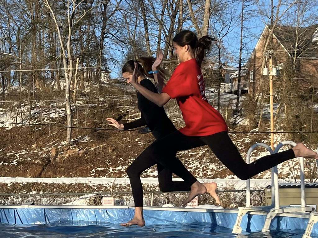 Two girls, one dressed in all black and the other wearing black pants and a red shirt, have just jumped off the steps of an above ground pool. Neither girl has entered the water yet. The left foot of the girl dressed in all back is just about to break the surface of the water.