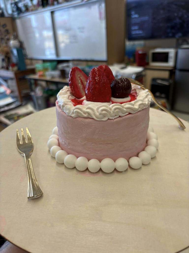 Art sculpture made to look like a strawberry cake. At the base of the cake sculpture are white balls. On top of the cake sculpture are strawberry sculptures. There is also a fork to the left of the cake and a fork leaning against the cake. 