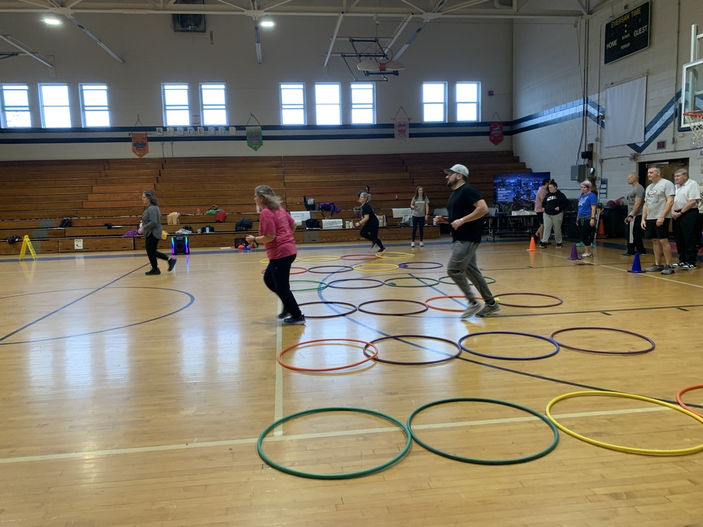 There are six, evenly spaced out, rows of hula hoops on a gym floor with four hula hoops in each row. Teachers are taking turns running in and out of the hula hoops.