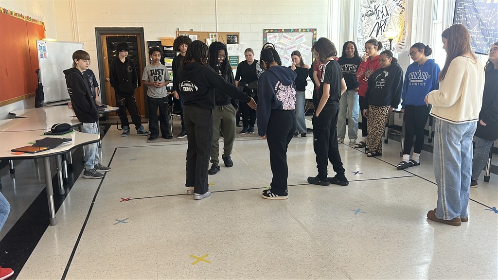A life size graph has been made out of tape on the floor of a classroom. Students are standing at different spots on the graph. The students are acting as data points on the graph as part of a math lesson. 