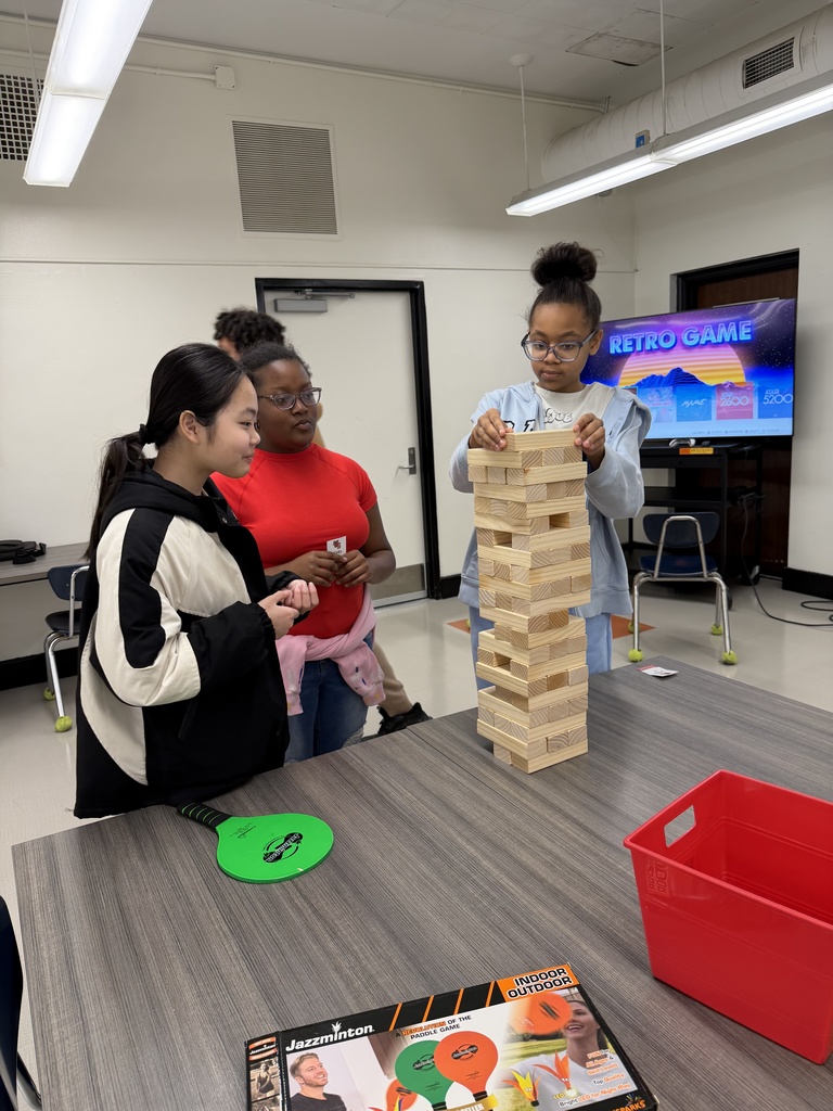 Three students are standing around a table playing jenga. Two students are watching as the third student is placing one of the wooden blocks on top of the stack of blocks. 