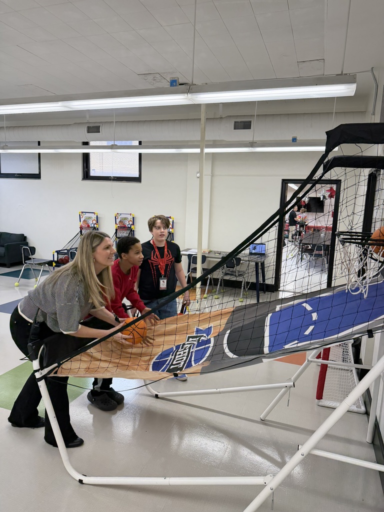 The school principal is playing arcade like mini basketball game with a student trying to see who can make more baskets in a period of time. A third student is standing to the side of the game watching. 