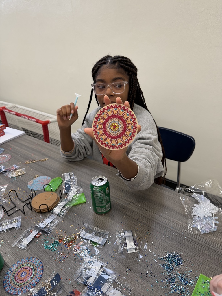 A student sitting at an arts and crafts table that is covered in glitter. She is holding up a dream catcher that she designed with glitter and beads.