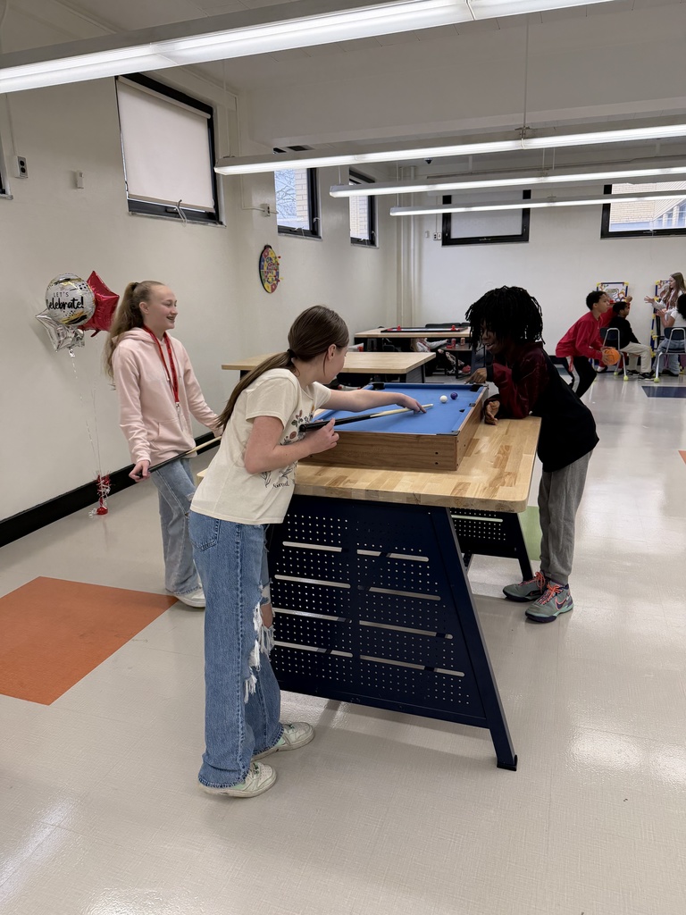 Two students are playing a game of pool on a mini pool table. A third student standing to the left of the pool table, is also holding a pool cue and smiling while one student lines up for a shot.