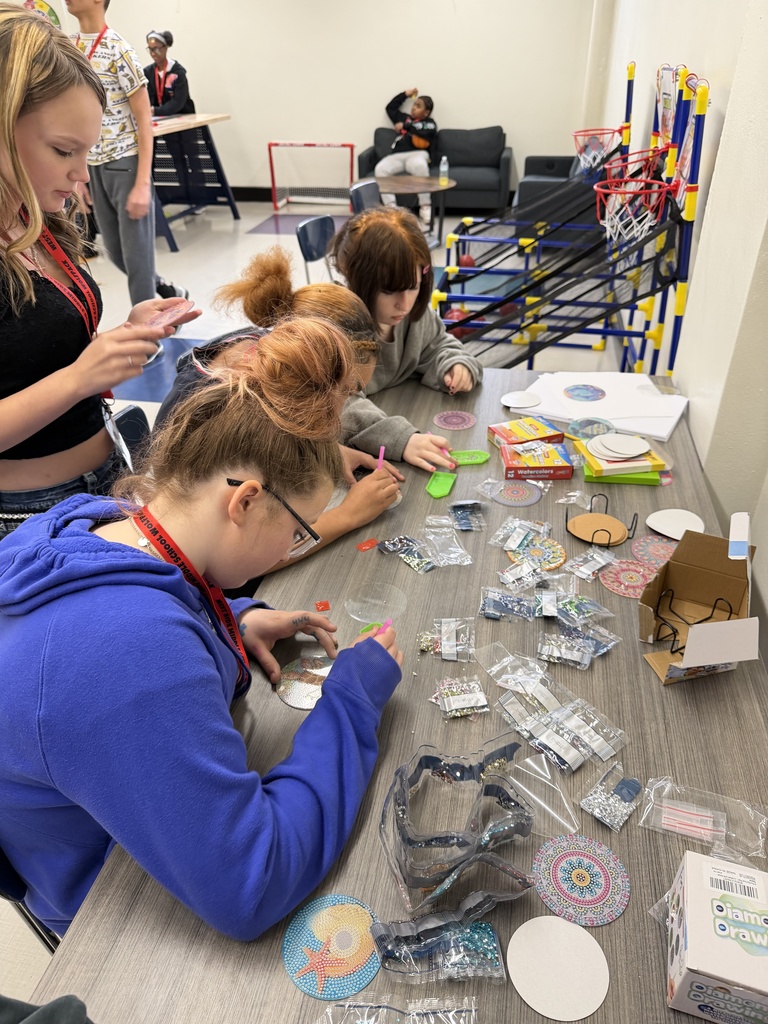 Three students sitting at an arts and crafts table, and one student standing up behind the three sitting down. They are working on an art project and there are beads and glitter covering the table. 