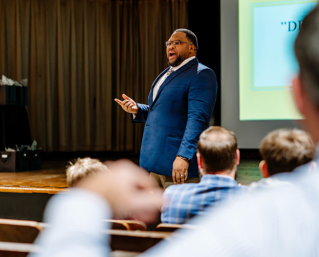 Dr. Brian Dinkins is standing in front of the stage in the Dunbar Middle School Auditorium speaking to students and their mentors. 