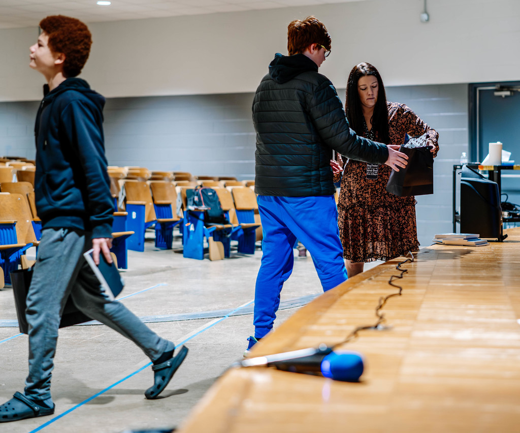 The school principal is handing a student his bag with that has his dress shirt and tie in it. Another student, who has already received his bag, is walking back towards his seat. 