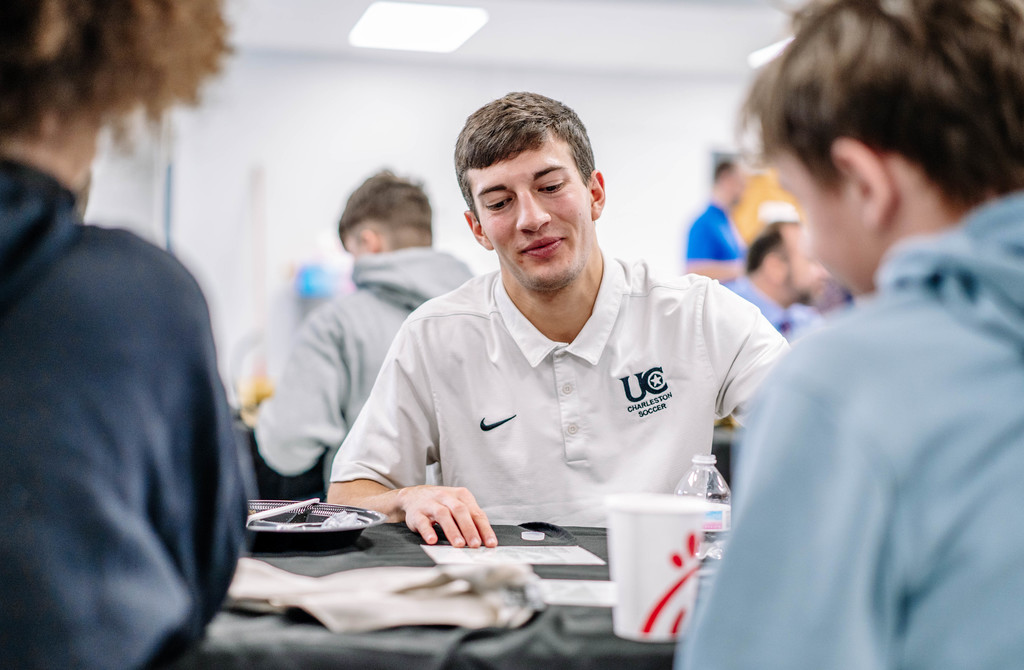 A University of Charleston soccer player is speaking with two students at the Guys in Ties event while eating breakfast.