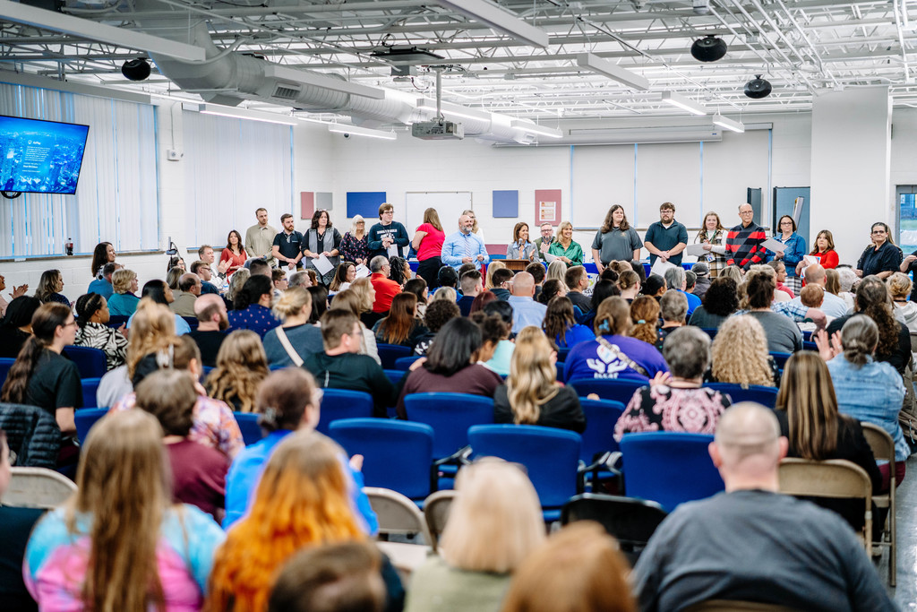 A wide shot of the board meeting where teachers were recognized. The picture is taken from the back of the room, behind all of the audience seating. Most of the seats are full. In front of the audience, the teachers are lined up. In the middle of the teachers is KCS Superintendent, Paula Potter, Asst. Superintendent of Secondary Gene Smith, and Deputy Superintendent, Missy Ruddle.