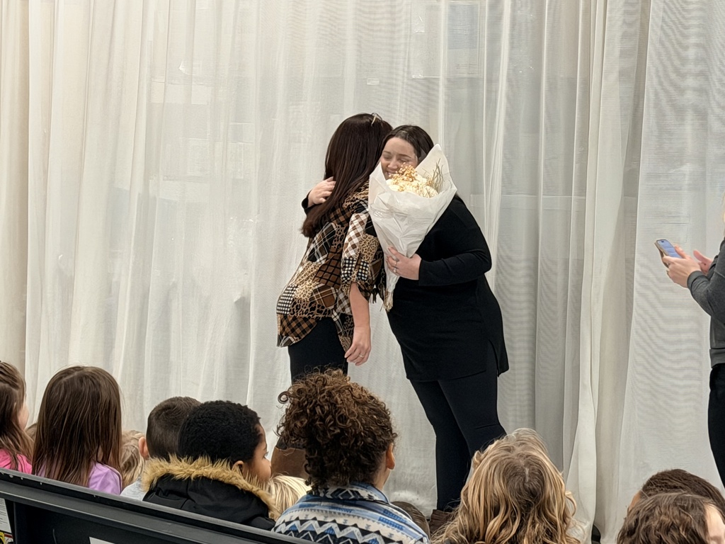 Taylor is hugging her school's principal. Taylor is holding a bouquet of flowers in her left hand. Students sitting in the audience are looking on .