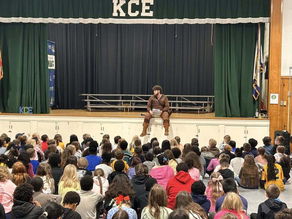 The WVU mascot is sitting on the stage at the school holding a book. Students are sitting on the floor in front of the stage. 