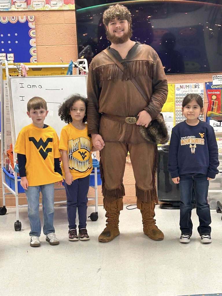 The WVU mascot is standing with three students. He is holding the hand of the student to his right. Two students are wearing gold WVU shirts and the other is wearing a blue WVU sweatshirt. 