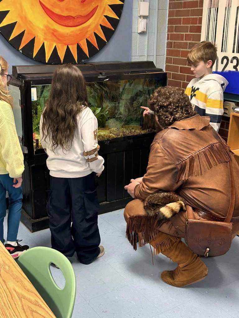 The WVU mascot is squatting down looking at a fish tank in a classroom. To the right of him, a student is pointing at the one of the fish in the tank. Two students are standing to the left of the mascot also looking at the fish tank.