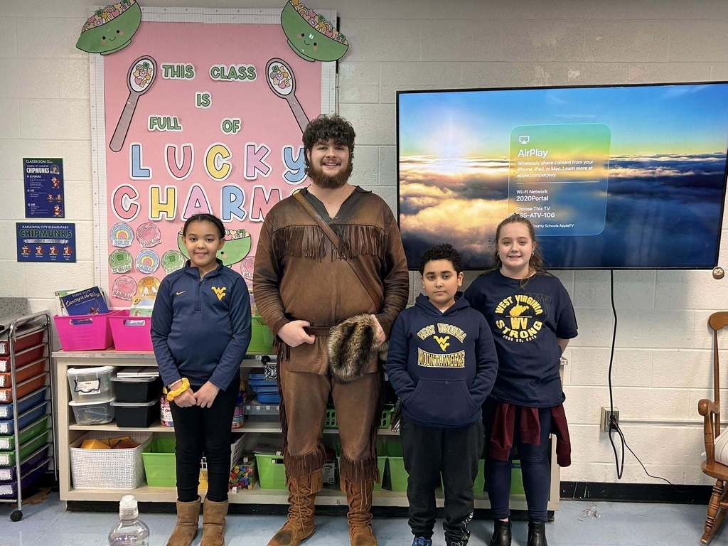 The WVU mascot is posing for a picture with three students in a classroom. All three students are wearing blue WVU gear.