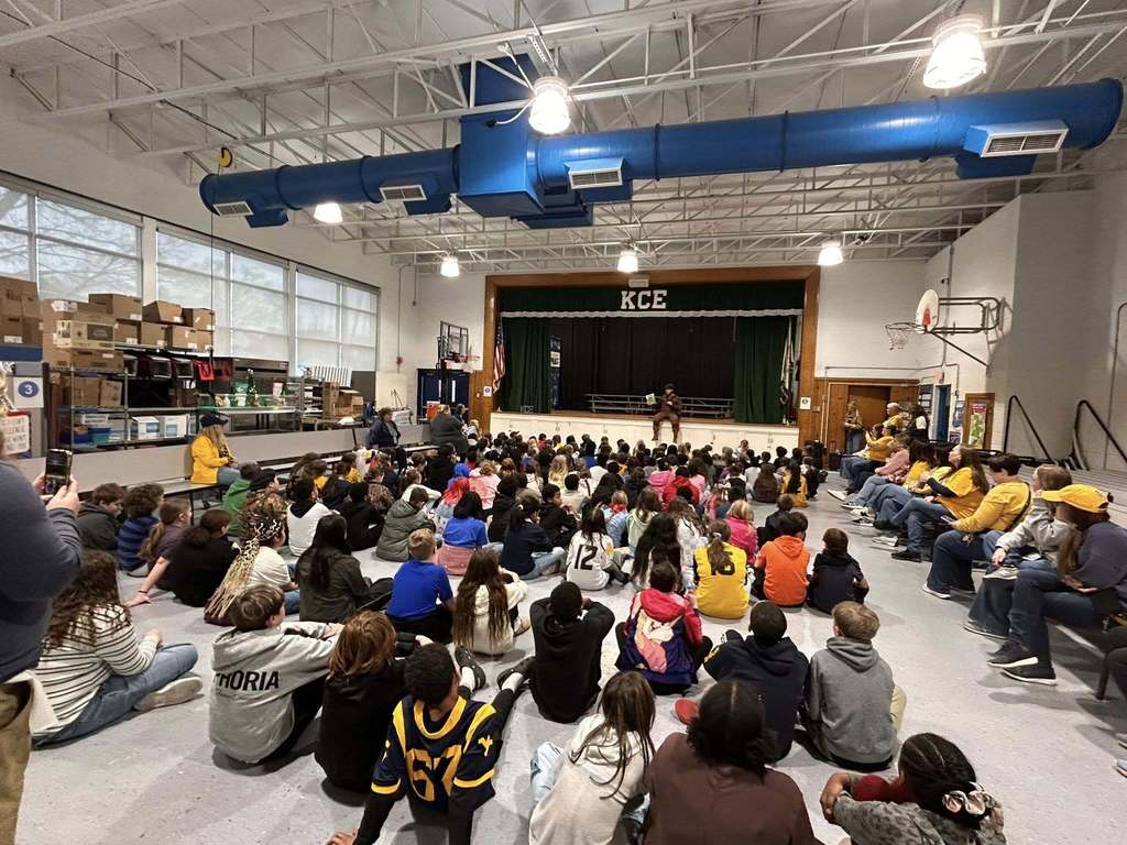 The WVU mascot is sitting on the stage reading a book to students. In front of the stage, students are sitting on the floor and on the side school staff are sitting on benches as the Mountaineer reads. 
