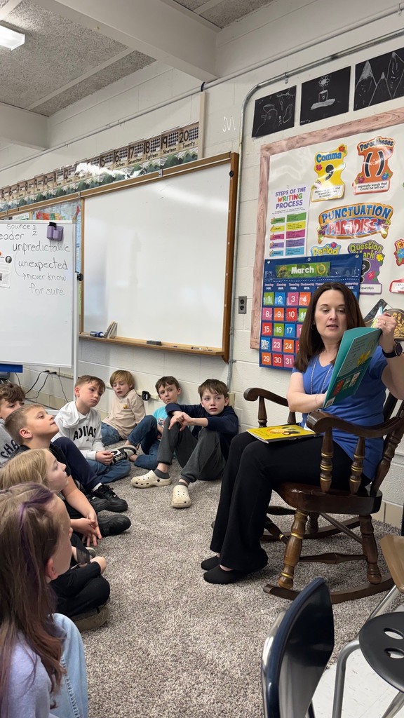 KCS Treasure, Melanie Meadows, is sitting in a wooden rocking chair in the front of a classroom. She is reading a Dr. Seuss book to students, who are seated on the carpet in front of her. Melanie is holding the book to her side so student's can see the pictures. 