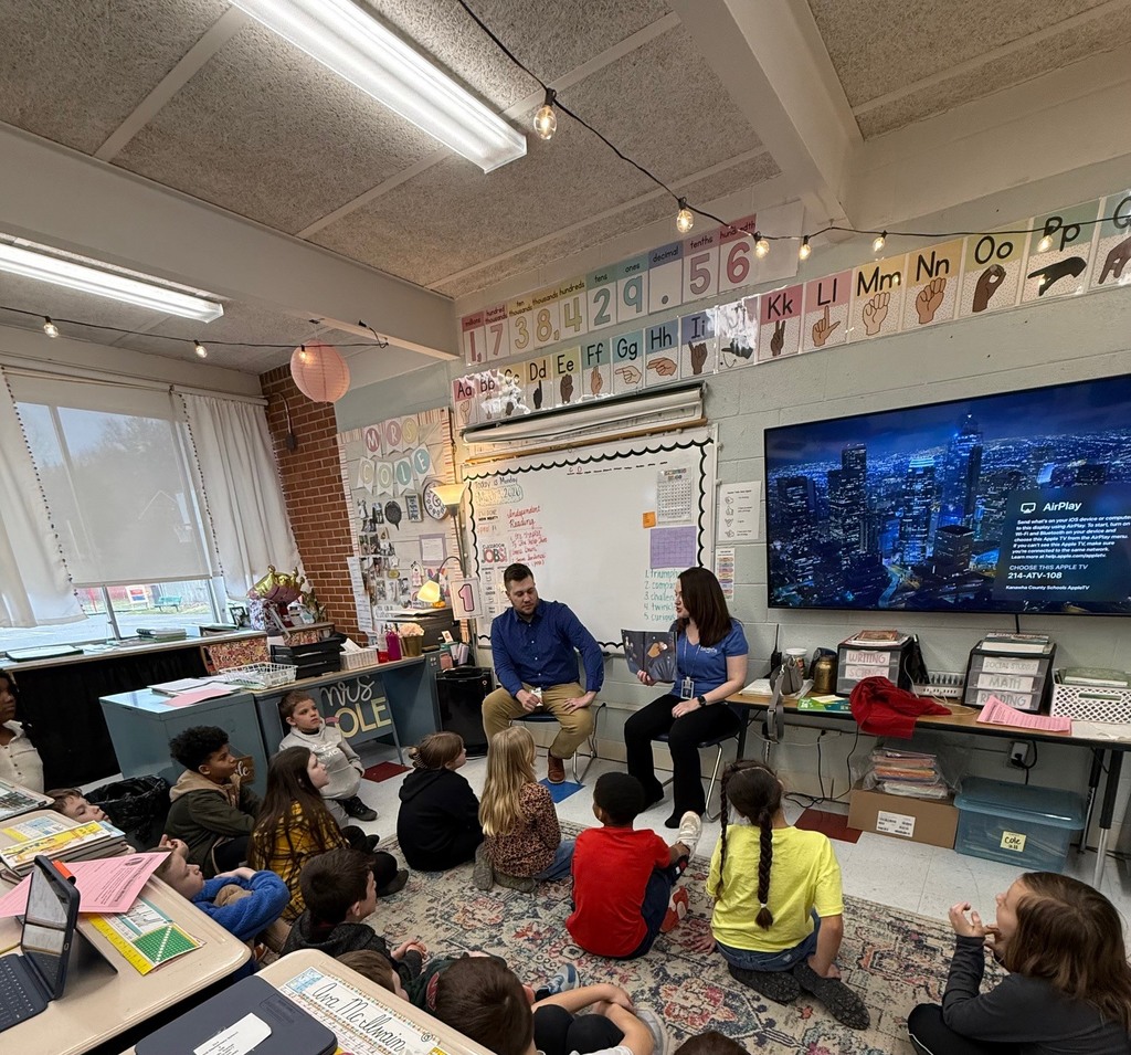 KCS Treasurer, Melanie Meadows, and Purchasing Director, Josh Lewis, are sitting at the front of a classroom. Melanie is holding a book about Jane Goodall. Students are sitting on a rug in front of Josh and Melanie. 