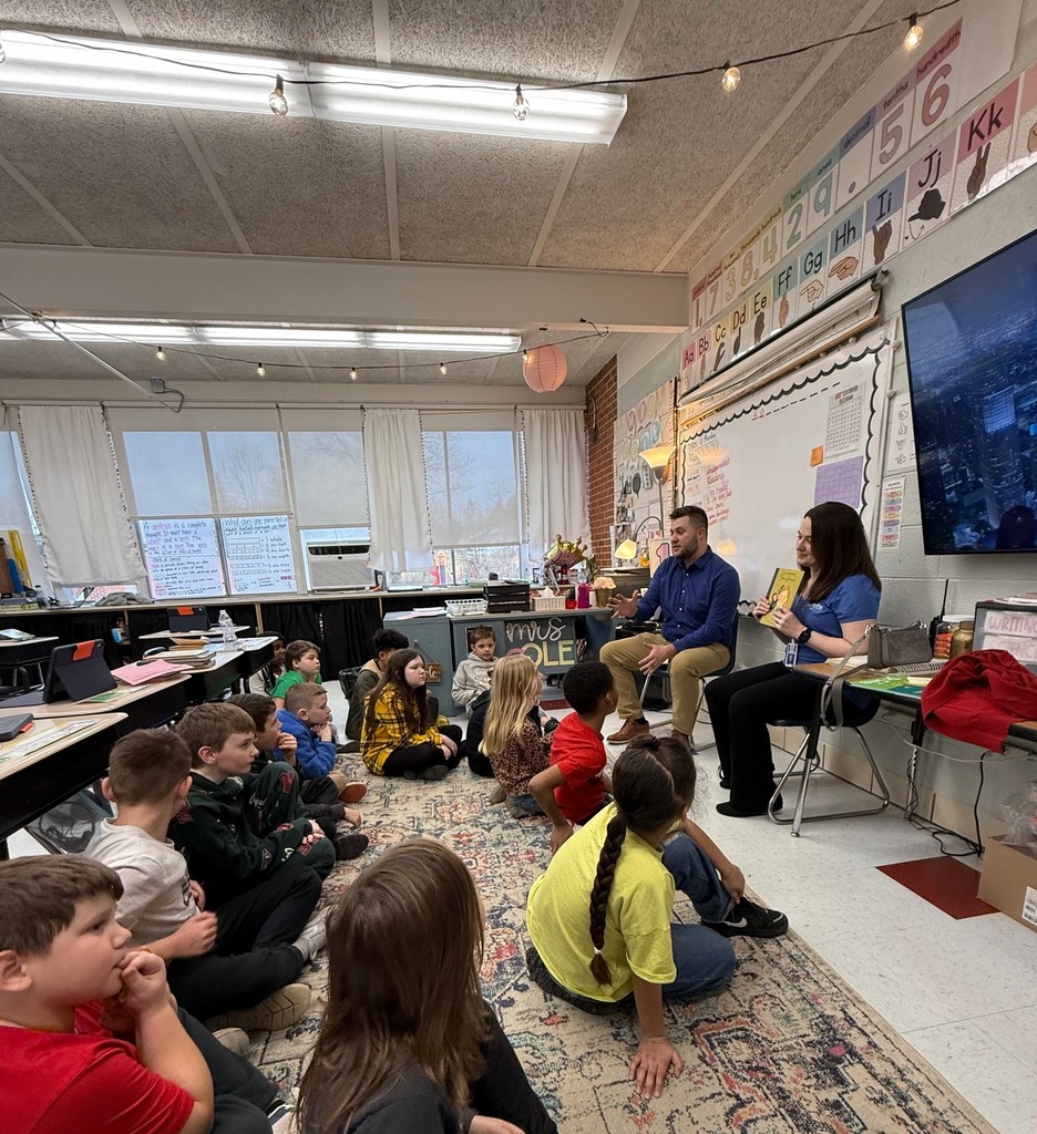 KCS Treasurer, Melanie Meadows, and Purchasing Director, Josh Lewis, are sitting at the front of a classroom. Melanie is holding a book about Jane Goodall. Students are sitting on a rug in front of Josh and Melanie. 