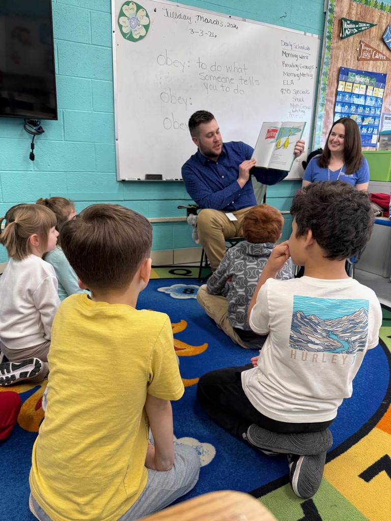 KCS Treasurer, Melanie Meadows, and Purchasing Director, Josh Lewis, are sitting at the front of a classroom. Josh is reading a Dr. Seuss book. Students are sitting on a rug in front of Josh and Melanie. 