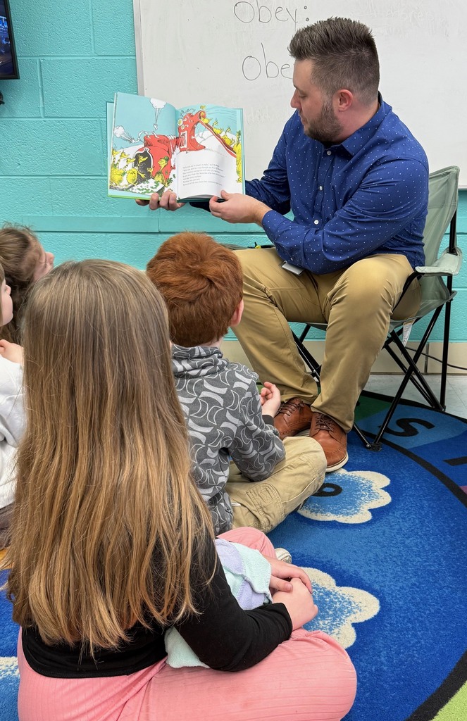 KCS Purchasing Director, Josh Lewis, is sitting in a soccer style folding chair. He is reading a Dr. Seuss book to students, who are seated on the carpet in front of him. Josh is holding the book to his side so student's can see the pictures. 