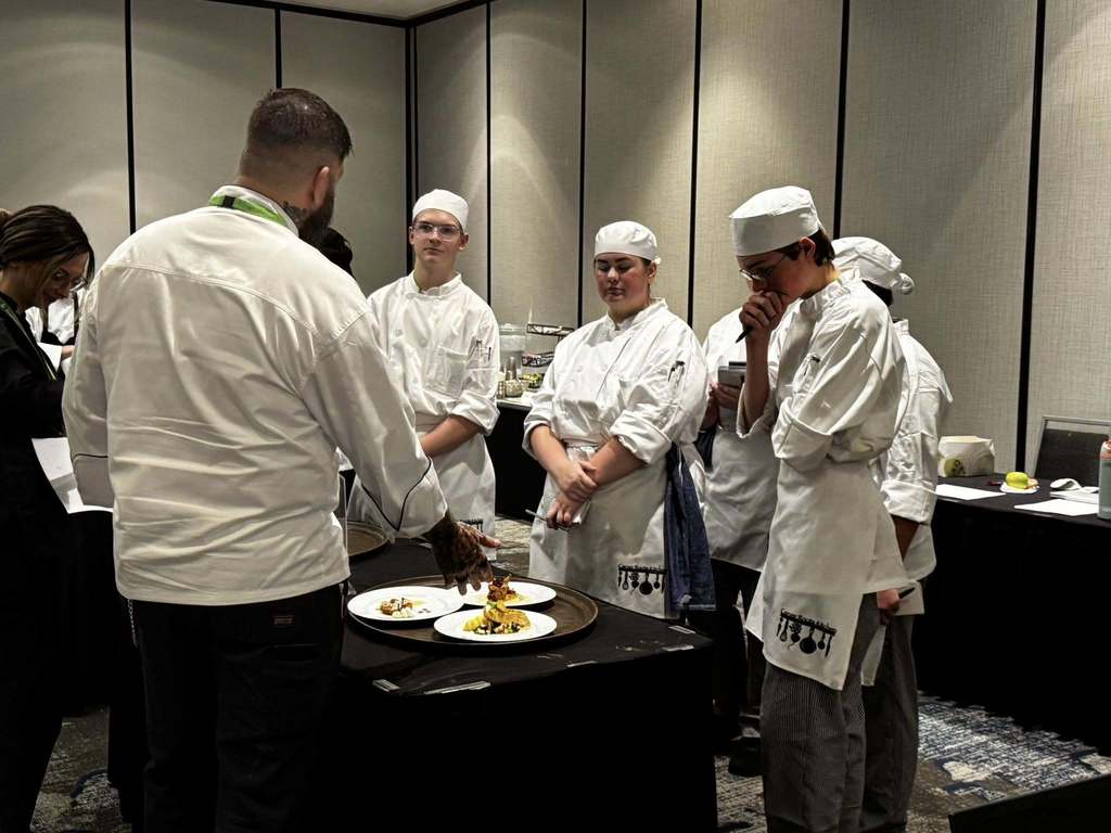Five culinary students, dressed in their chefs outfits, are standing behind their three course meal. A professional chef is on the other side of the table and is gesturing to one of the dishes. 