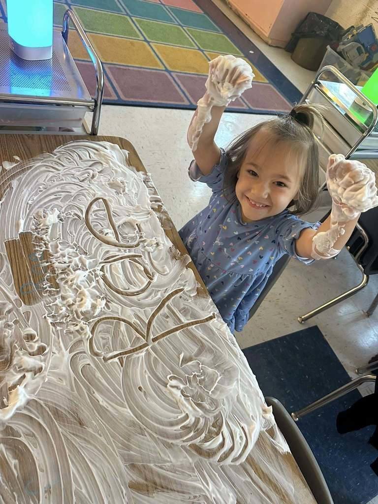 A young student sitting at her school table, smiling at the camera, and has both of her arms in the air. Her arms and table are covered in shaving cream that was used for a writing lesson.