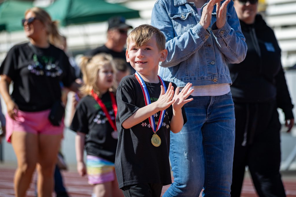 A young student is clapping his hands. He is wearing a gold medal around his neck. The medal has the number 1 on it. Behind him adults are clapping and other students are wearing medals. 