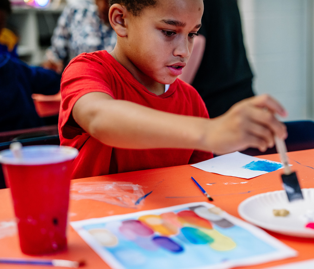 Student sitting at an art table. He is painting a white sheet of paper that is sitting in front of him. He is dipping his sponge brush into paint that has been squirted on to a paper plate.